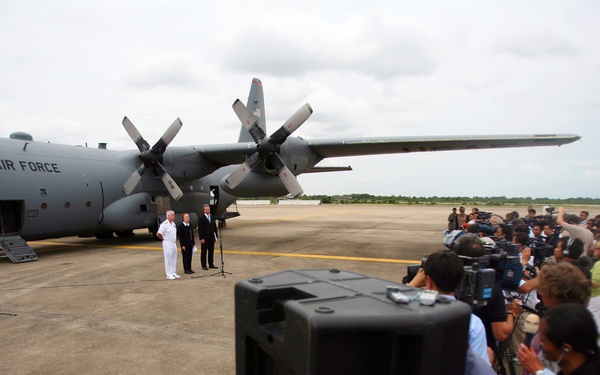 A press conference or official statement is being delivered on an airport tarmac in front of a U.S. Air Force C-130 Hercules transport aircraft. Several officials, including one in a white naval uniform, stand at a microphone podium while a large group of journalists and camera operators record the event. The scene suggests a significant military or diplomatic occasion drawing substantial media attention.