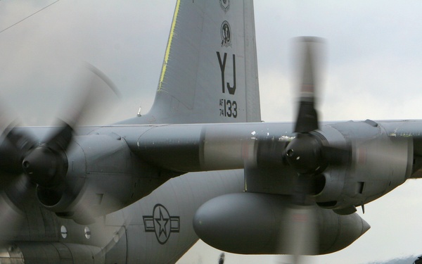 A U.S. Air Force C-130 Hercules transport aircraft is shown on a flight line with its four turboprop engines running, propellers visibly spinning in motion blur. A ground crew member in a flight suit and headset stands behind the aircraft, monitoring the engine run-up. A second C-130 is visible in the background.