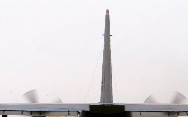 A military transport aircraft, consistent with a C-130 Hercules, is photographed head-on from the rear on an active taxiway or runway. All four turboprop engines are running with propellers in motion, visible as blurred discs. A second aircraft and a small guard post or checkpoint structure are visible in the background.