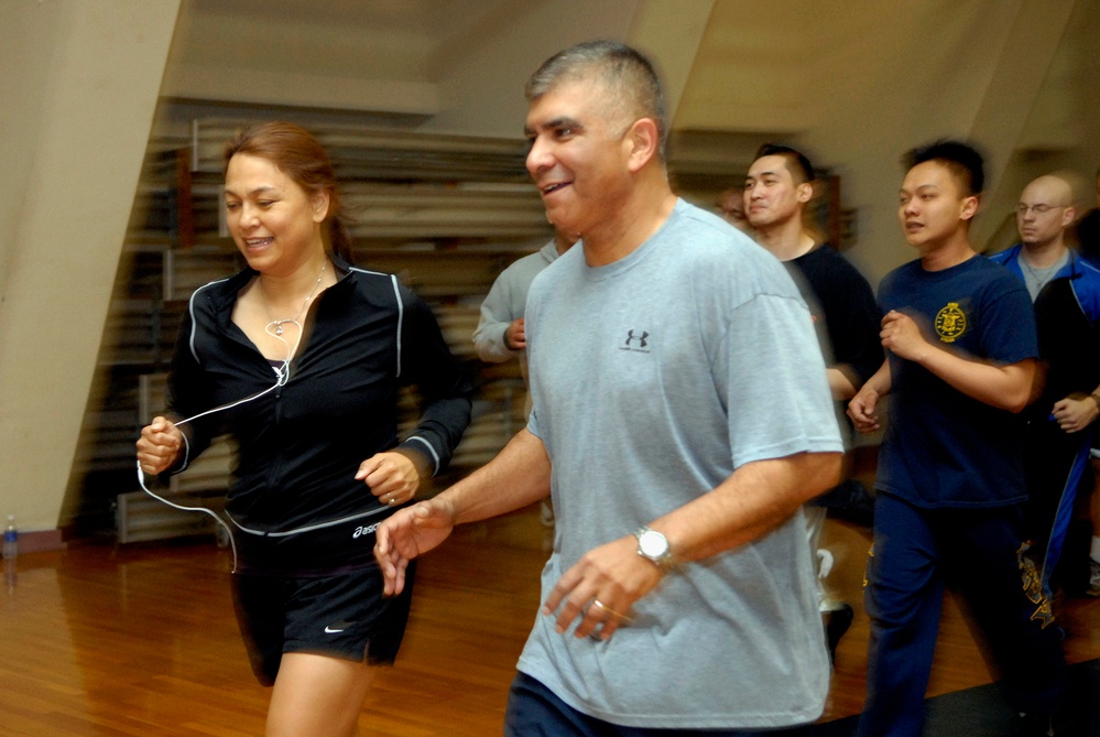 A group of people are jogging or running together inside what appears to be a gymnasium with a hardwood floor. Two individuals are prominently featured in the foreground — a woman in a black Asics tracksuit with earphones and a man in a gray Under Armour t-shirt wearing a watch. Several other participants are visible in the background, suggesting a group fitness or physical training session.