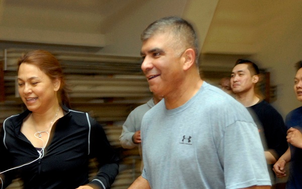 A group of people are jogging or running together inside what appears to be a gymnasium with a hardwood floor. Two individuals are prominently featured in the foreground — a woman in a black Asics tracksuit with earphones and a man in a gray Under Armour t-shirt wearing a watch. Several other participants are visible in the background, suggesting a group fitness or physical training session.