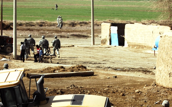 Military personnel in combat uniforms are gathered around a yellow CAT (Caterpillar) bulldozer in what appears to be a rural Middle Eastern or Central Asian setting. Several soldiers are inspecting or working on the construction equipment while others stand guard in the background near civilians and additional military personnel. The scene suggests a military engineering or civil affairs operation near mud-brick structures and agricultural fields.