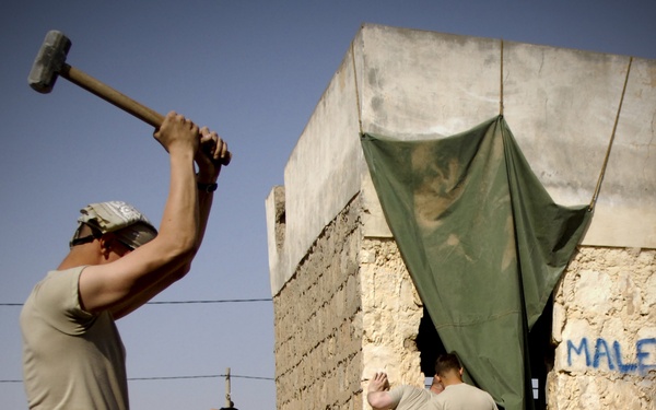 Military personnel in U.S. Army combat uniforms are engaged in construction or demolition work at a rudimentary stone/concrete structure. One soldier in the foreground is swinging a large sledgehammer, while others work near a doorway covered by a green tarp with the word 'MALE' painted on the wall. The setting appears to be a forward operating base or field installation in an arid environment.