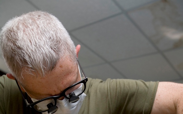 A male dental professional with gray hair is performing a dental procedure on a reclined patient. The practitioner is wearing magnifying loupes attached to black-framed glasses, a surgical mask, latex gloves, and an olive green t-shirt. He is using dental instruments inside the patient's open mouth.