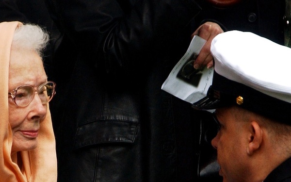 A uniformed military service member wearing a white-topped dress cap and white gloves is presenting a folded American flag to an elderly woman wearing an orange/peach head scarf and glasses. This appears to be a military funeral or memorial ceremony, a solemn occasion where the flag is traditionally presented to the next of kin of a deceased service member. Several other individuals are visible in the background, some holding what appears to be a printed program or photograph.