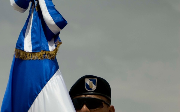 A soldier in desert camouflage uniform holds a blue and white flag with gold fringe, identifiable as the flag of El Salvador based on visible text reading 'AMERICA CENTR' and the coat of arms. The soldier wears a black beret with a shield insignia and dark sunglasses. A second similarly uniformed soldier is visible in the background, suggesting a formal military color guard ceremony.