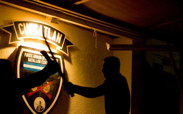 A nighttime photograph showing two silhouetted figures in front of an illuminated military insignia on a wall. The emblem reads 'CUSCATLAN' at the top and 'EL SALVADOR' at the bottom, with text indicating 'Fuerza Armada de El Salvador, Batallon Cuscatlan'. The dramatic backlighting creates strong silhouettes of the two individuals, one of whom appears to be holding a long object such as a staff or weapon.