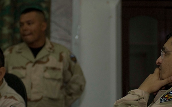 Three military personnel are gathered in what appears to be an indoor briefing or meeting setting. Two individuals are seated at a table with documents and a water bottle visible, while a third stands in the background. All three wear desert camouflage uniforms with shoulder patches.
