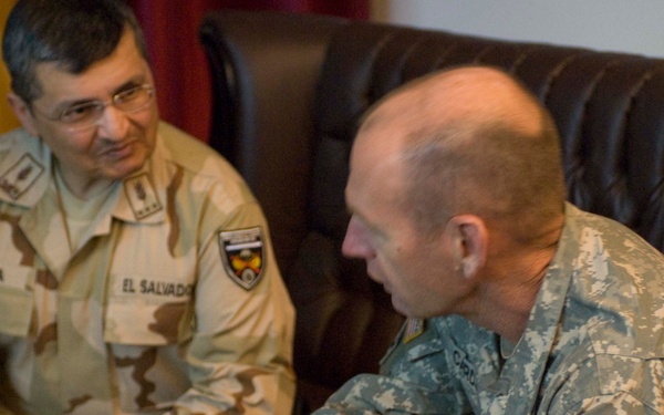Two military personnel are seated and engaged in conversation. The soldier on the left wears a desert camouflage uniform with 'EL SALVADOR' and a partial surname 'OLINA' visible on name tapes, along with a unit patch. The soldier on the right wears US Army ACU (Army Combat Uniform) with a 3rd Infantry Division patch on the left sleeve.