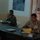 Two military personnel in camouflage uniforms are seated at a dark conference table in what appears to be a briefing or meeting room. An Iraqi flag is visible on the left wall, and a map or aerial photograph is posted on the wall behind them. One individual appears to be speaking while the other reviews a yellow document.