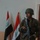 A soldier in camouflage uniform and green helmet stands at attention inside a building, holding an assault rifle. Two Iraqi flags are displayed on stands behind him against a light-colored wall. The setting appears to be an official or administrative interior space.