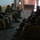 Several armed soldiers in military fatigues and helmets are resting on floral-patterned sofas inside a building. They are holding rifles and wearing combat gear including body armor and boots. A sixth figure is visible standing in a doorway or hallway in the background.