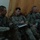 Three U.S. Army soldiers in full combat gear are seated together, likely during a briefing or meeting in an indoor setting. The soldier in the center is writing notes on a clipboard or notepad. All soldiers are wearing ACU (Army Combat Uniform) pattern camouflage and are equipped with weapons, body armor, and tactical gear.