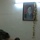 A uniformed military officer sits at a desk in what appears to be an office or command post. Behind him on the wall hangs a framed portrait of a religious figure with Arabic text beneath it, decorated with artificial flowers. The setting suggests a military or paramilitary administrative space.