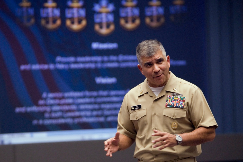 A U.S. Navy officer in khaki service uniform is presenting or speaking in front of a projected slide displaying Navy insignia (anchors) and text including 'Mission' and 'Vision' sections. The officer wears multiple service ribbons, a name tag reading 'CAMPA', and additional qualification badges on the left chest. The setting appears to be a formal military briefing or conference room.