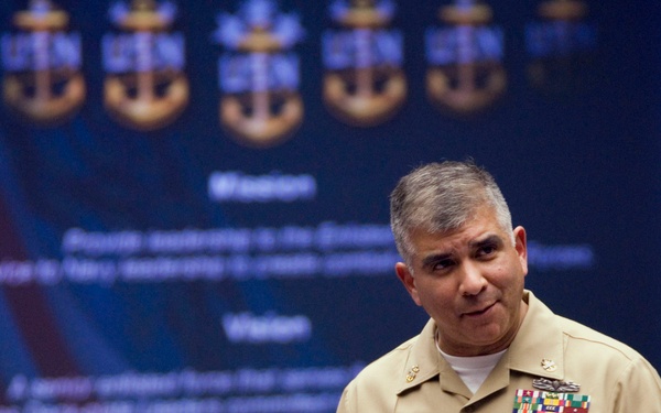 A U.S. Navy officer in khaki service uniform is presenting or speaking in front of a projected slide displaying Navy insignia (anchors) and text including 'Mission' and 'Vision' sections. The officer wears multiple service ribbons, a name tag reading 'CAMPA', and additional qualification badges on the left chest. The setting appears to be a formal military briefing or conference room.