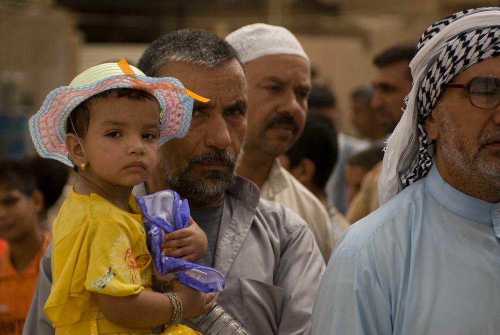 A group of men and a young child are gathered together in what appears to be a public or communal setting. A man holds a small child wearing a colorful sun hat and yellow shirt, who is clutching a purple plastic bag. The individuals are dressed in traditional Middle Eastern attire, suggesting a cultural or religious gathering.
