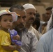 A group of men and a young child are gathered together in what appears to be a public or communal setting. A man holds a small child wearing a colorful sun hat and yellow shirt, who is clutching a purple plastic bag. The individuals are dressed in traditional Middle Eastern attire, suggesting a cultural or religious gathering.