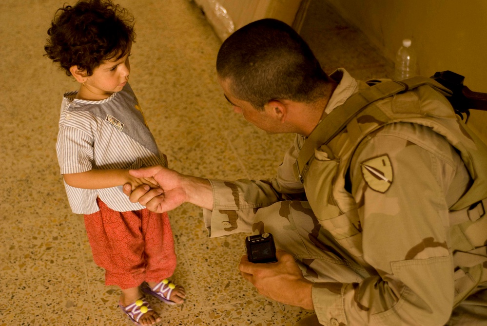 A soldier in desert camouflage uniform crouches down to interact with a young child, gently holding the child's hand. The soldier wears a unit patch on the left shoulder and holds a small black device in the other hand. The setting appears to be an indoor tiled floor environment.