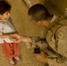 A soldier in desert camouflage uniform crouches down to interact with a young child, gently holding the child's hand. The soldier wears a unit patch on the left shoulder and holds a small black device in the other hand. The setting appears to be an indoor tiled floor environment.