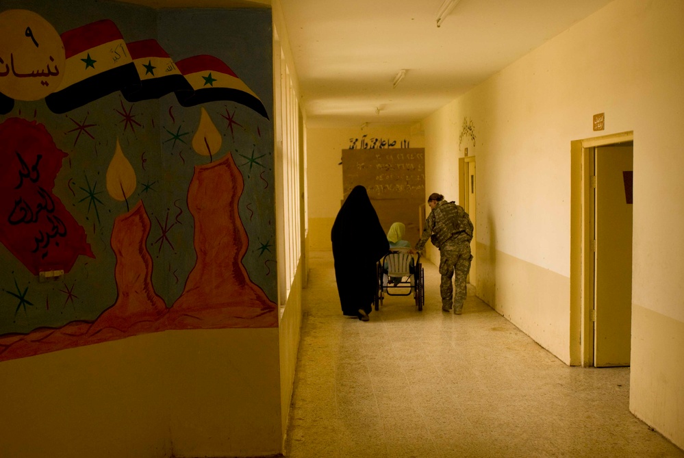 A dimly lit institutional corridor, likely a hospital in Iraq, showing a woman in a black abaya and a soldier in military fatigues assisting a person in a wheelchair. The left wall features a colorful mural with the Iraqi flag, candles, and Arabic text including the word 'Nisan' (April). The scene suggests a humanitarian or medical assistance context.
