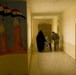 A dimly lit institutional corridor, likely a hospital in Iraq, showing a woman in a black abaya and a soldier in military fatigues assisting a person in a wheelchair. The left wall features a colorful mural with the Iraqi flag, candles, and Arabic text including the word 'Nisan' (April). The scene suggests a humanitarian or medical assistance context.
