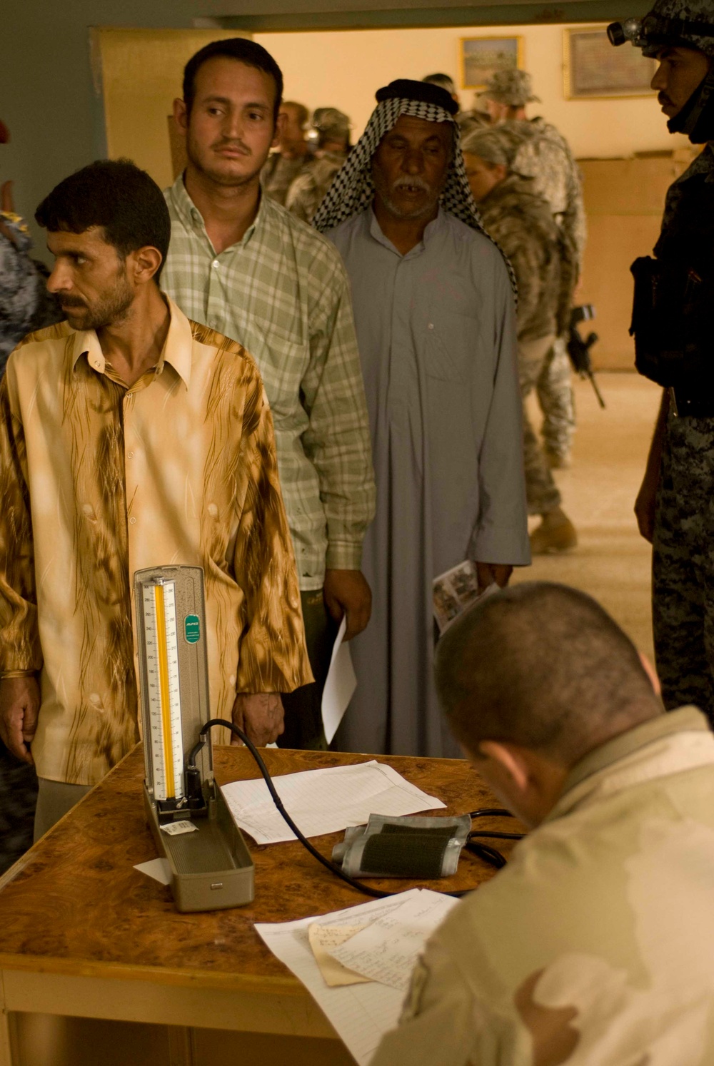 The image shows a medical screening or civil affairs event where civilian men are queued in a line at a table staffed by a uniformed personnel. A sphygmomanometer (blood pressure cuff and gauge) is visible on the table along with paperwork, suggesting a medical assessment is being conducted. Military personnel in combat uniforms are visible in the background providing security or oversight.