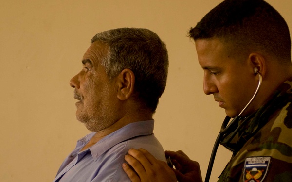 A person in military camouflage uniform is using a stethoscope to examine an older civilian man's back or chest area. The medical examination appears to be taking place against a plain beige/tan wall. The uniformed individual has a shoulder patch visible on their left arm, suggesting an official military or paramilitary medical role.