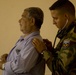 A person in military camouflage uniform is using a stethoscope to examine an older civilian man's back or chest area. The medical examination appears to be taking place against a plain beige/tan wall. The uniformed individual has a shoulder patch visible on their left arm, suggesting an official military or paramilitary medical role.