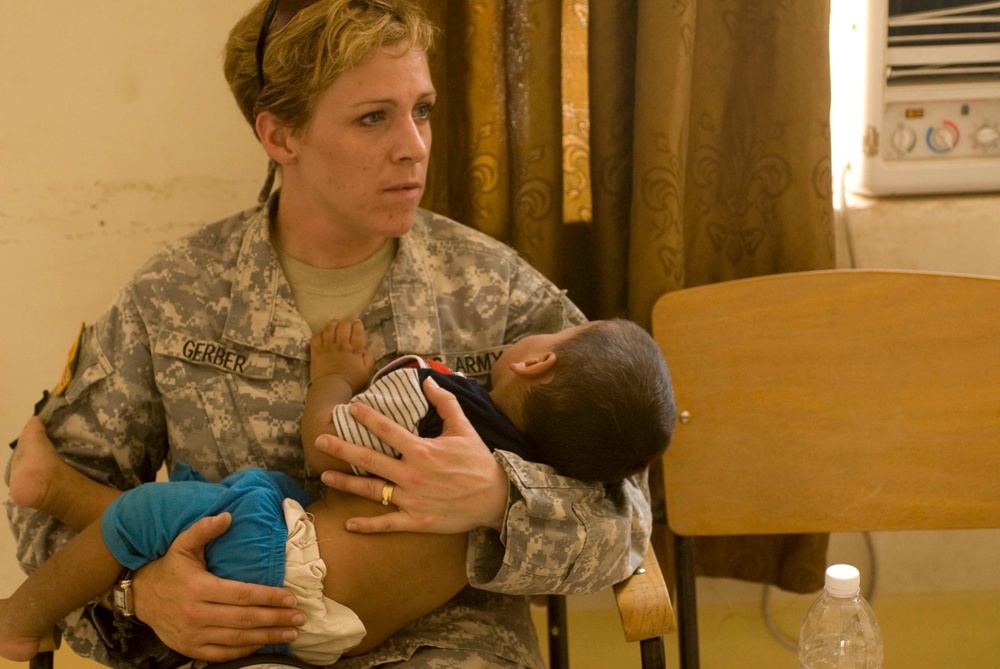 A U.S. Army soldier identified by the name tape 'GERBER' is seated and holding a young infant in her arms. She is wearing the Army Combat Uniform (ACU) pattern and appears to be in an indoor setting, possibly a medical or community outreach facility. The scene suggests a humanitarian or civil affairs mission.