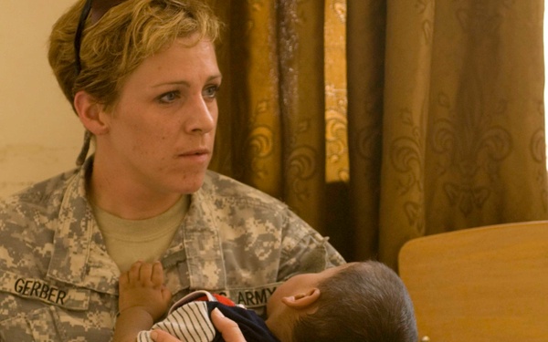 A U.S. Army soldier identified by the name tape 'GERBER' is seated and holding a young infant in her arms. She is wearing the Army Combat Uniform (ACU) pattern and appears to be in an indoor setting, possibly a medical or community outreach facility. The scene suggests a humanitarian or civil affairs mission.