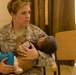 A U.S. Army soldier identified by the name tape 'GERBER' is seated and holding a young infant in her arms. She is wearing the Army Combat Uniform (ACU) pattern and appears to be in an indoor setting, possibly a medical or community outreach facility. The scene suggests a humanitarian or civil affairs mission.