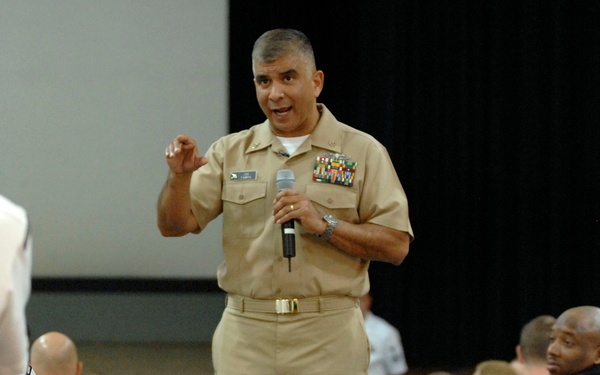 A senior military officer in tan/khaki service uniform is addressing a large audience of sailors dressed in white uniforms, seated in pews or auditorium-style seating. The speaker is holding a microphone and gesturing with one hand, suggesting an active speech or briefing. The setting appears to be a military auditorium or chapel used for an all-hands or formal assembly.