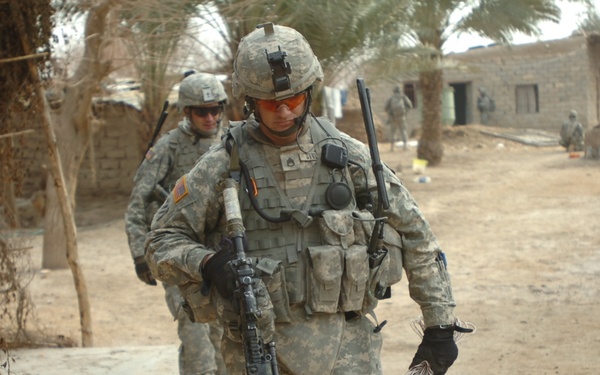 The image shows U.S. Army soldiers in Universal Camouflage Pattern (UCP) uniforms conducting a patrol or operation in what appears to be a desert village environment. The foreground soldier is fully equipped with body armor, helmet, rifle, radio equipment, and is carrying a coil of rope or cord. The setting suggests a forward operating area in a Middle Eastern or Central Asian theater of operations.