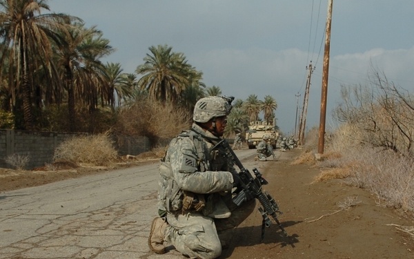 The image shows U.S. military soldiers in combat gear taking up positions along a cracked road flanked by palm trees and dry brush. A primary soldier in the foreground is kneeling on the road's edge holding an assault rifle, while additional soldiers and at least one military vehicle are visible in the background. The setting appears to be a rural road in a Middle Eastern environment.