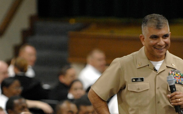 A senior U.S. Navy officer in khaki service uniform is addressing a large audience of military personnel in what appears to be an auditorium or assembly hall. He is holding a microphone and smiling, suggesting an informal or motivational speaking engagement. The audience, dressed in both white and dark uniforms, appears engaged and amused.