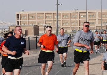 A large group of U.S. Army personnel are running on an outdoor track at what appears to be a military installation, likely in a Middle Eastern or arid environment based on the architecture and landscape. The runners are wearing a mix of Army PT (physical training) gear including gray 'ARMY' shirts, black shorts, and yellow reflective belts. One individual sits near a concrete barrier on the right side of the track, wearing a military backpack.