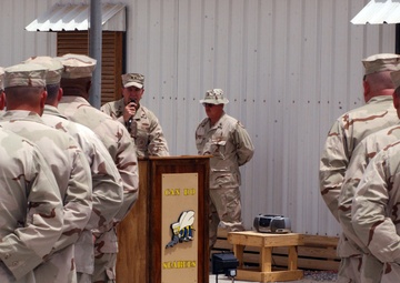A military ceremony or briefing is taking place outdoors at what appears to be a forward operating base. A speaker addresses a formation of personnel from behind a wooden podium bearing the 'Can Do Seabees' insignia, indicating this is a U.S. Navy Construction Battalion (Seabees) unit. Personnel are wearing desert camouflage uniforms (DCU - three-color desert pattern) consistent with operations in the Middle East.