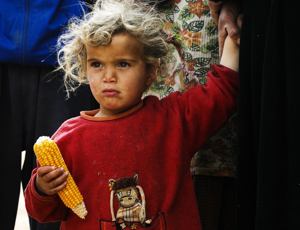 A young child with curly blonde hair and a solemn expression holds a partially eaten ear of corn. The child is wearing a red fleece sweatshirt with an animal graphic and appears to be standing near adults. The child's clothing shows signs of dirt and wear, and their expression conveys a serious or distressed demeanor.