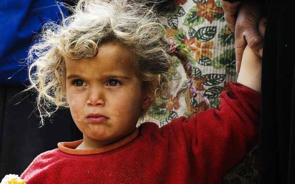 A young child with curly blonde hair and a solemn expression holds a partially eaten ear of corn. The child is wearing a red fleece sweatshirt with an animal graphic and appears to be standing near adults. The child's clothing shows signs of dirt and wear, and their expression conveys a serious or distressed demeanor.