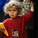A young child with curly blonde hair and a solemn expression holds a partially eaten ear of corn. The child is wearing a red fleece sweatshirt with an animal graphic and appears to be standing near adults. The child's clothing shows signs of dirt and wear, and their expression conveys a serious or distressed demeanor.