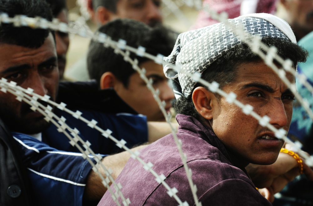 A group of people are gathered behind razor wire/concertina wire fencing. The foreground shows a young man wearing a traditional head covering and a purple jacket, looking sideways through the wire barrier. The scene suggests a crowd being held or restricted by a security barrier.