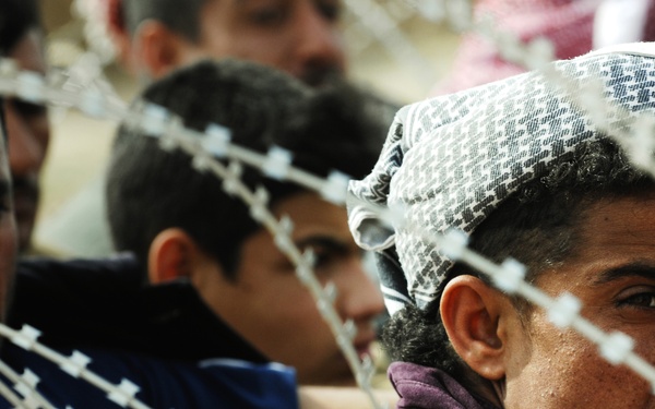 A group of people are gathered behind razor wire/concertina wire fencing. The foreground shows a young man wearing a traditional head covering and a purple jacket, looking sideways through the wire barrier. The scene suggests a crowd being held or restricted by a security barrier.