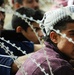 A group of people are gathered behind razor wire/concertina wire fencing. The foreground shows a young man wearing a traditional head covering and a purple jacket, looking sideways through the wire barrier. The scene suggests a crowd being held or restricted by a security barrier.