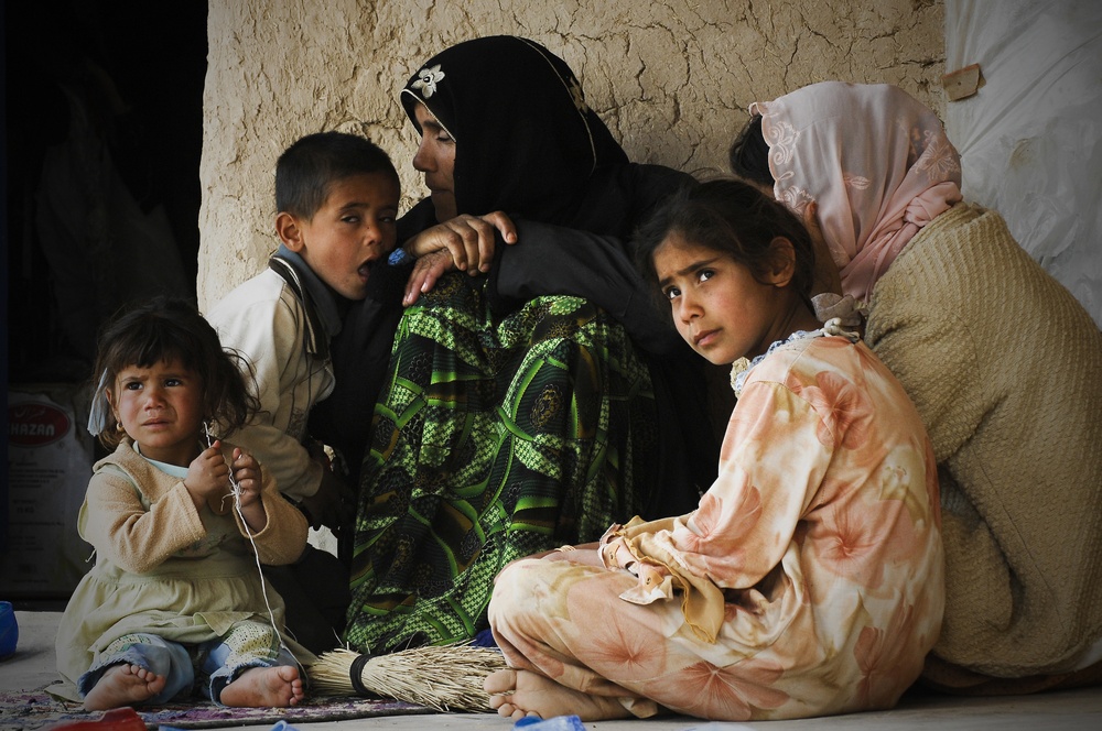 A group of women and young children are seated together on the floor against a cracked mud or clay wall. The scene suggests modest living conditions, with the individuals wearing traditional clothing including headscarves. The expressions on the children's faces convey distress or concern.