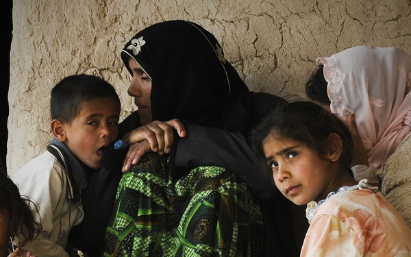 A group of women and young children are seated together on the floor against a cracked mud or clay wall. The scene suggests modest living conditions, with the individuals wearing traditional clothing including headscarves. The expressions on the children's faces convey distress or concern.