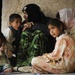 A group of women and young children are seated together on the floor against a cracked mud or clay wall. The scene suggests modest living conditions, with the individuals wearing traditional clothing including headscarves. The expressions on the children's faces convey distress or concern.