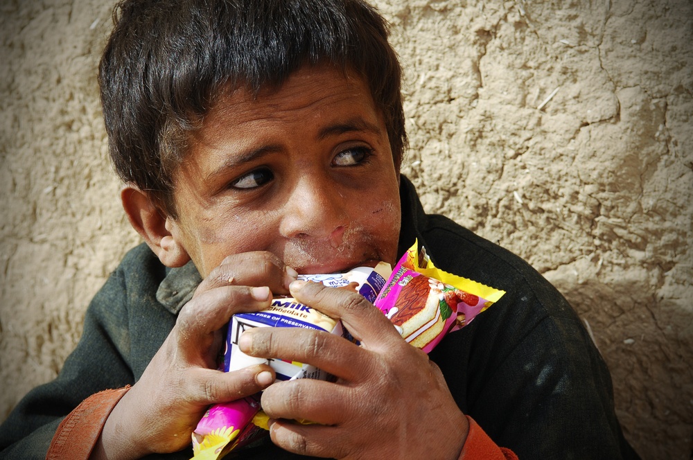 A young child is eating snack foods while seated against a rough mud or clay wall. The child is holding multiple packaged snack items simultaneously and appears to be eating eagerly. The setting and context suggest a modest or rural environment.