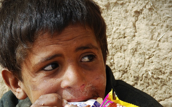 A young child is eating snack foods while seated against a rough mud or clay wall. The child is holding multiple packaged snack items simultaneously and appears to be eating eagerly. The setting and context suggest a modest or rural environment.