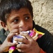 A young child is eating snack foods while seated against a rough mud or clay wall. The child is holding multiple packaged snack items simultaneously and appears to be eating eagerly. The setting and context suggest a modest or rural environment.
